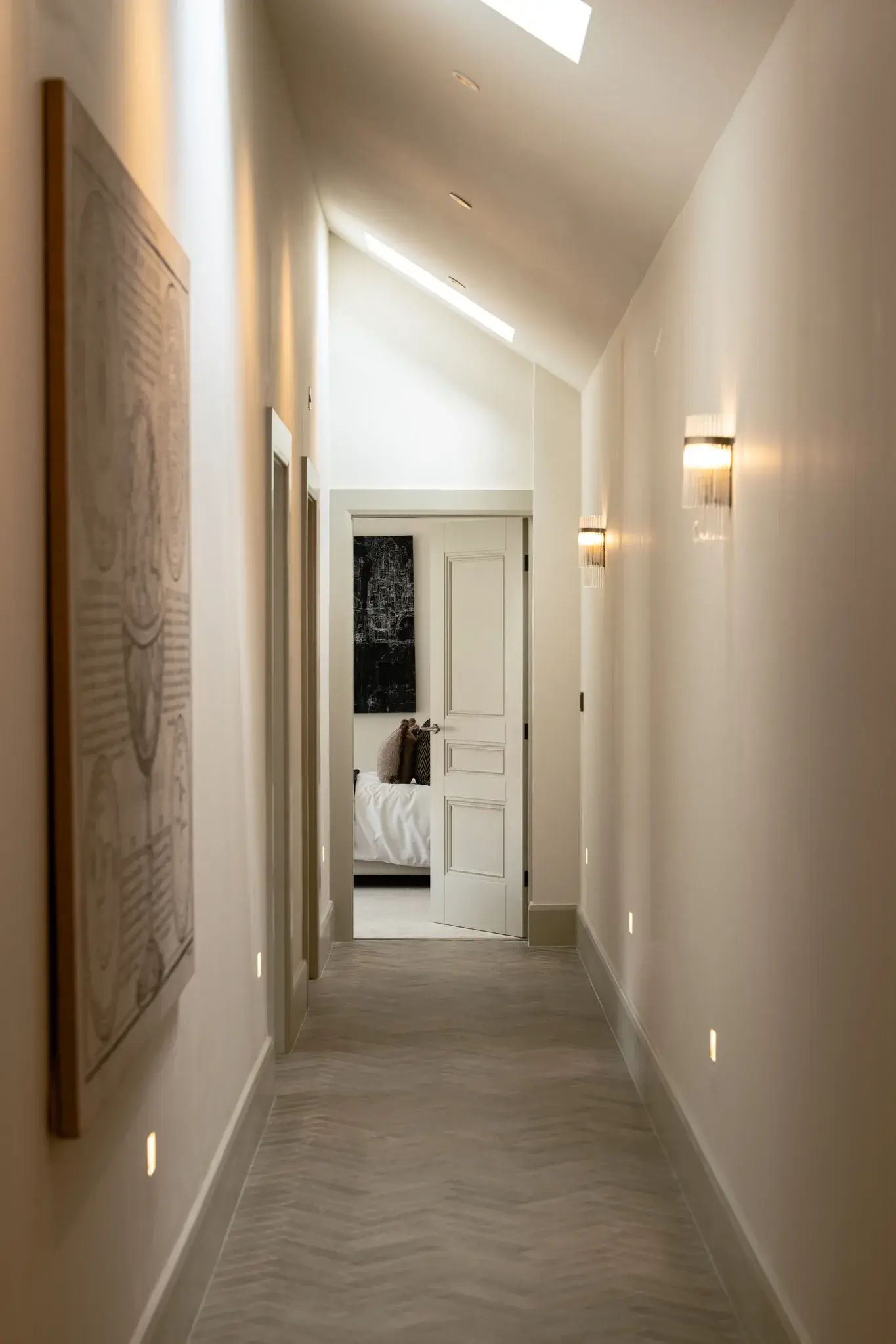 A hallway under a sloping ceiling with skylights, lit by wall lights and by small low-level lights set into the walls above the skirting