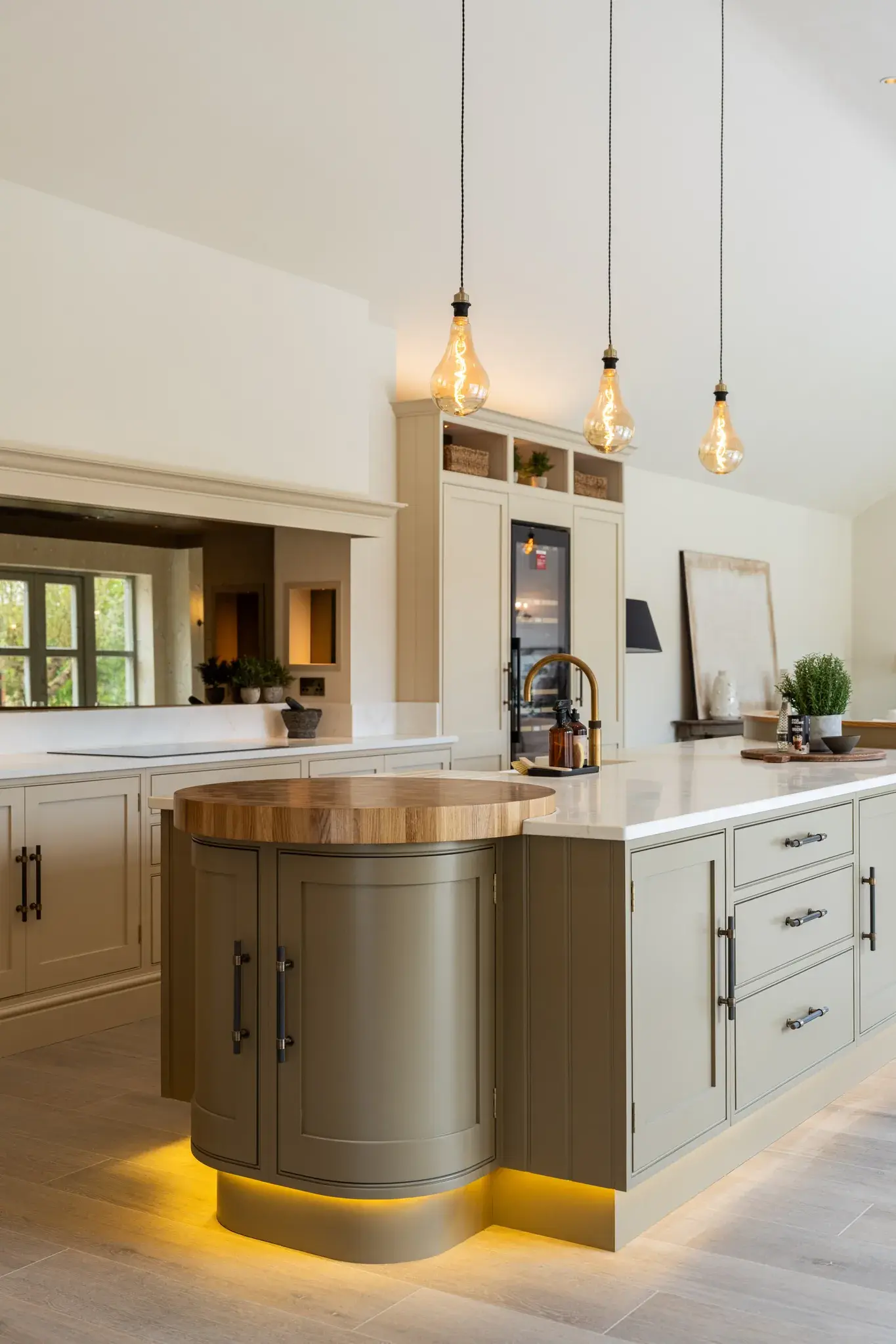 A kitchen island with a rounded wooden end, three filament pendant lights above it and a warm glow of light along its plinth