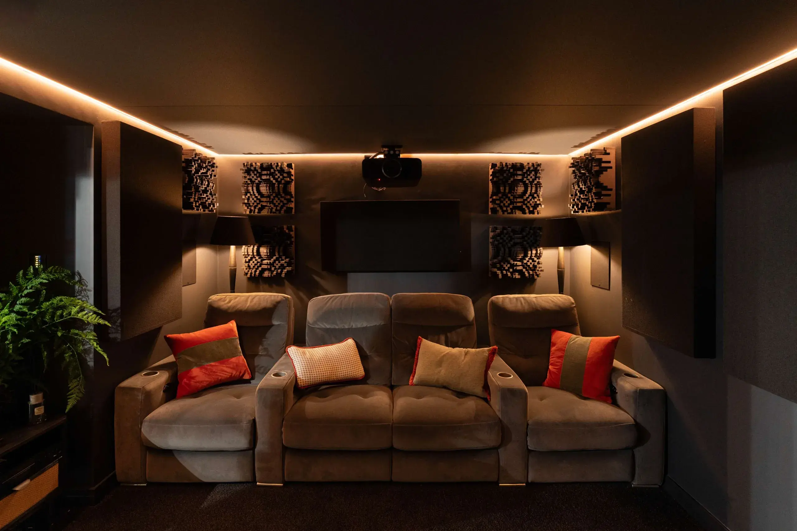 The showroom cinema room seen from the screen wall: suede recliner seats, patterned acoustic diffuser panels and a ceiling-mounted projector, with a warm light strip tracing the ceiling
